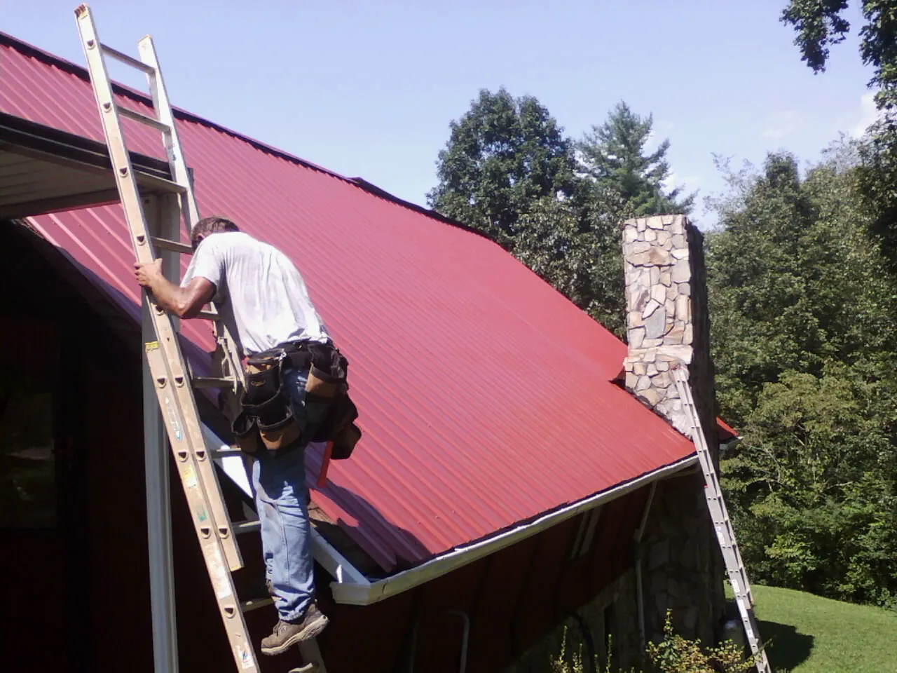 Roofer on a ladder beside a red metal roof and stone chimney in Shenandoah Junction